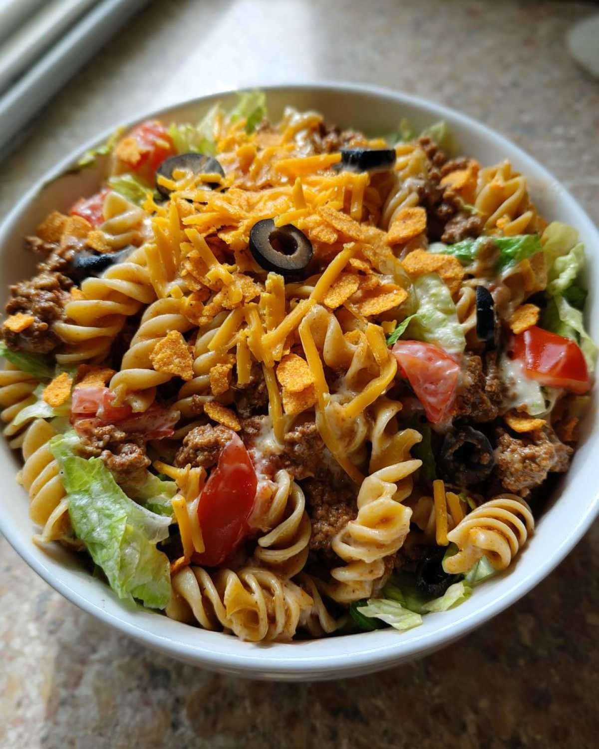 Overhead shot of dorito taco salad pasta in a white bowl, featuring pasta, lettuce, tomatoes, olives, and crushed Doritos.
