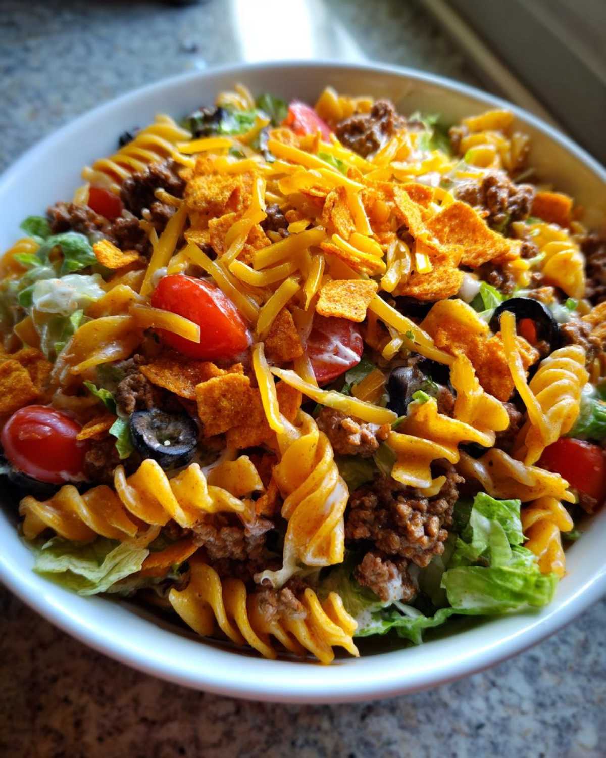 A bowl of Dorito taco salad pasta with lettuce, tomatoes, olives, ground beef, and crushed Doritos.