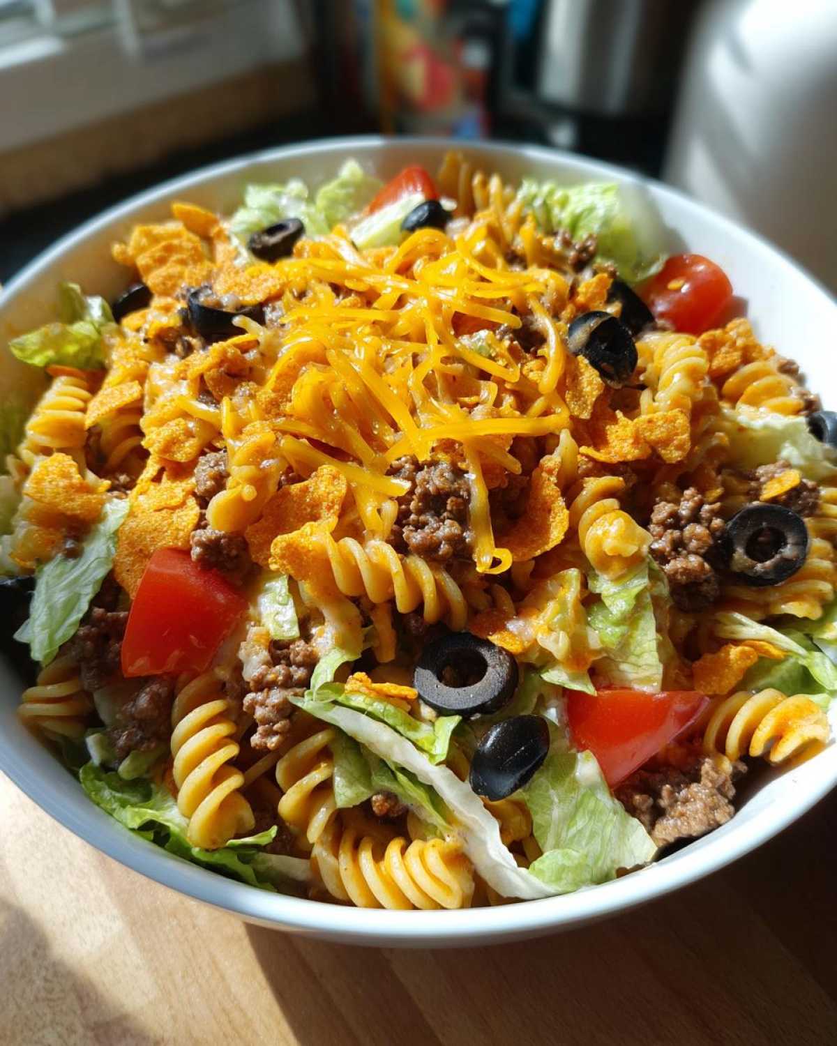 A bowl of Dorito taco salad pasta, featuring pasta, lettuce, tomatoes, olives, ground beef, cheese, and Doritos.