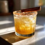 A delicious fall margarita in a glass with ice, a cinnamon-sugar rim, and a cinnamon stick garnish.