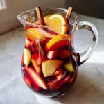 A glass pitcher filled with fall sangria, featuring sliced apples, oranges, pears, and cinnamon sticks.