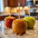 Three glossy caramel apples on sticks, one red and two green, sit on a speckled countertop.
