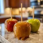 Three glossy caramel apples on sticks, one red and two green, sit on a speckled countertop.
