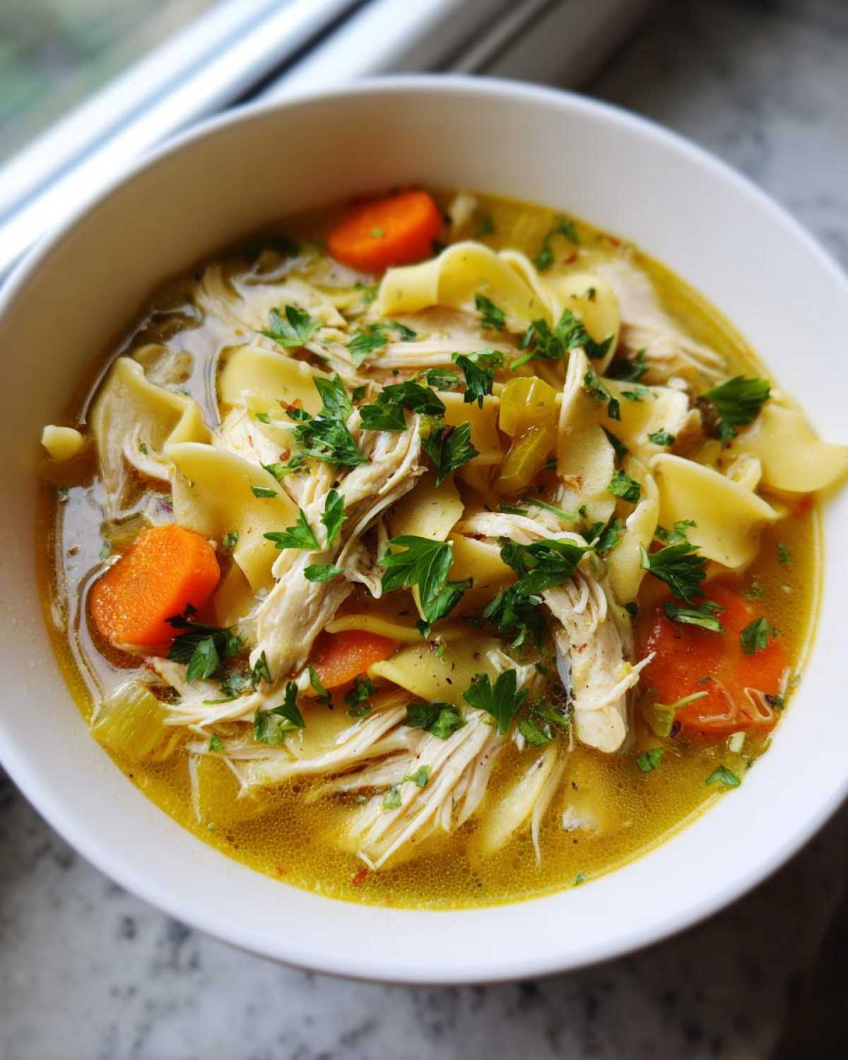 A close-up shot of a bowl of hearty chicken noodle soup, featuring shredded chicken, wide egg noodles, sliced carrots, and fresh parsley.
