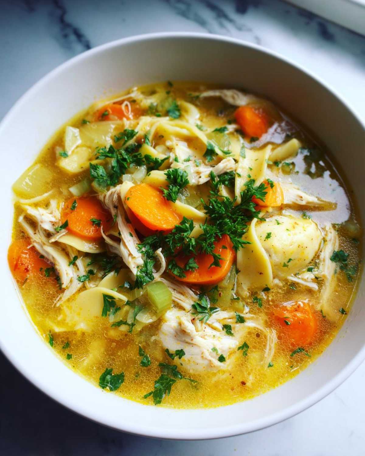 A close-up of a bowl of hearty chicken noodle soup, featuring shredded chicken, wide noodles, sliced carrots, and fresh parsley.