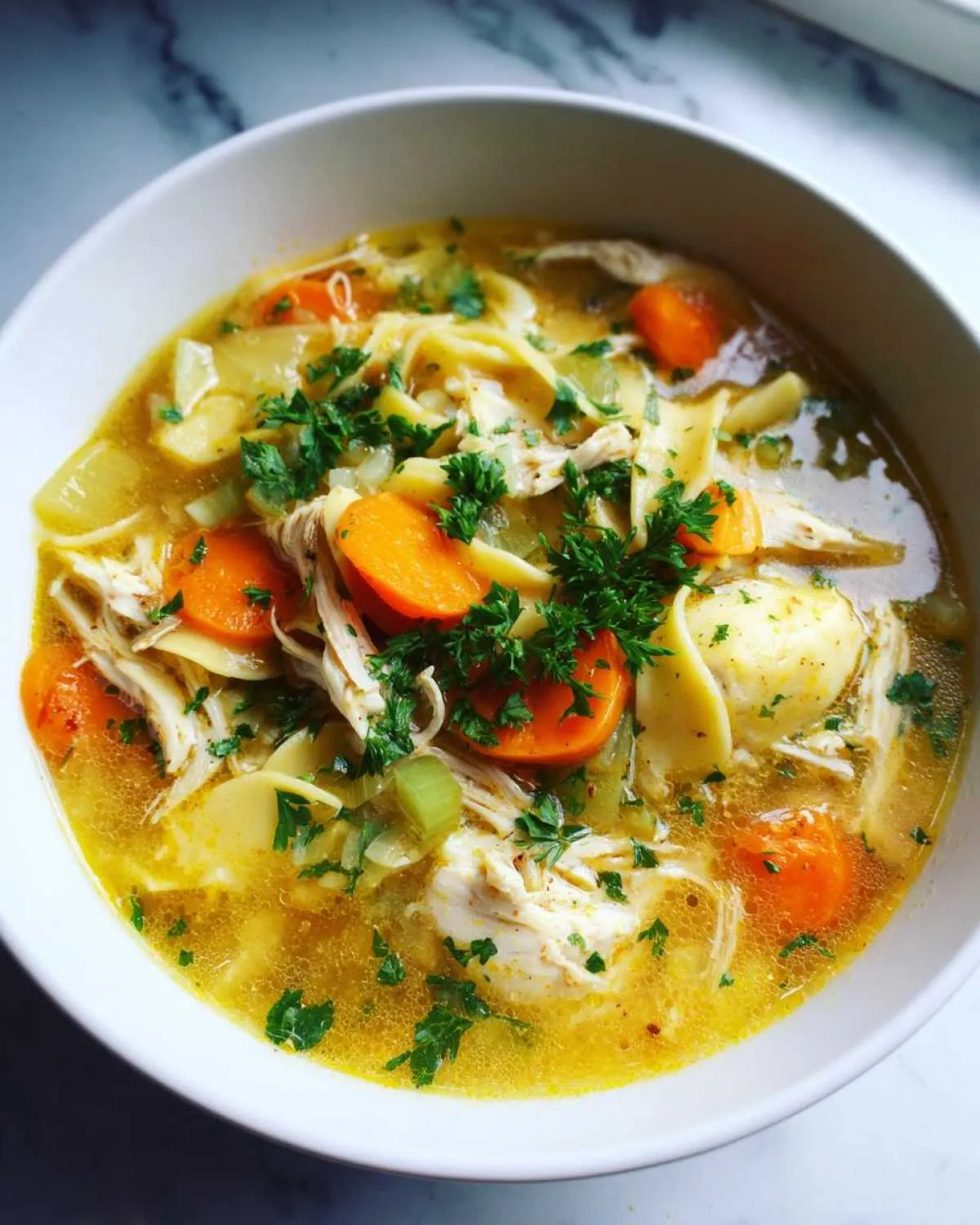 A close-up of a bowl of hearty chicken noodle soup, featuring shredded chicken, wide noodles, sliced carrots, and fresh parsley.