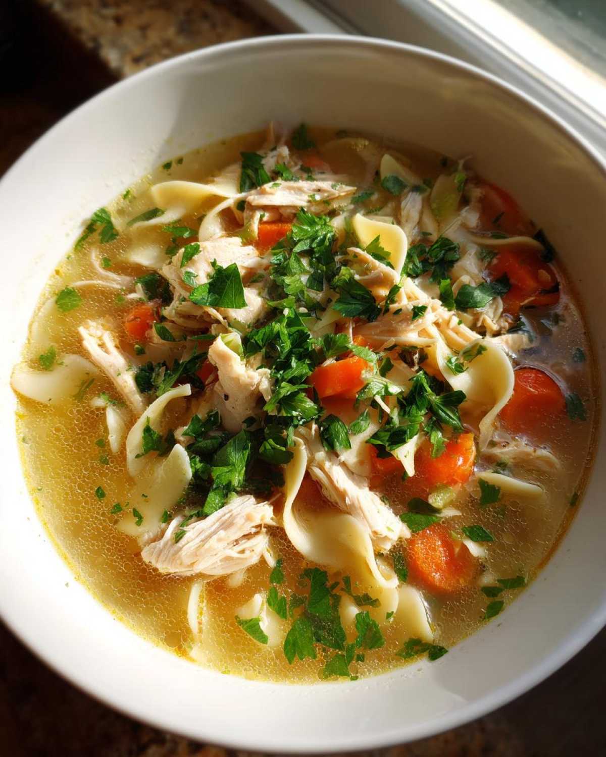 A close-up of a white bowl filled with hearty chicken noodle soup, featuring shredded chicken, wide noodles, carrots, and fresh parsley.
