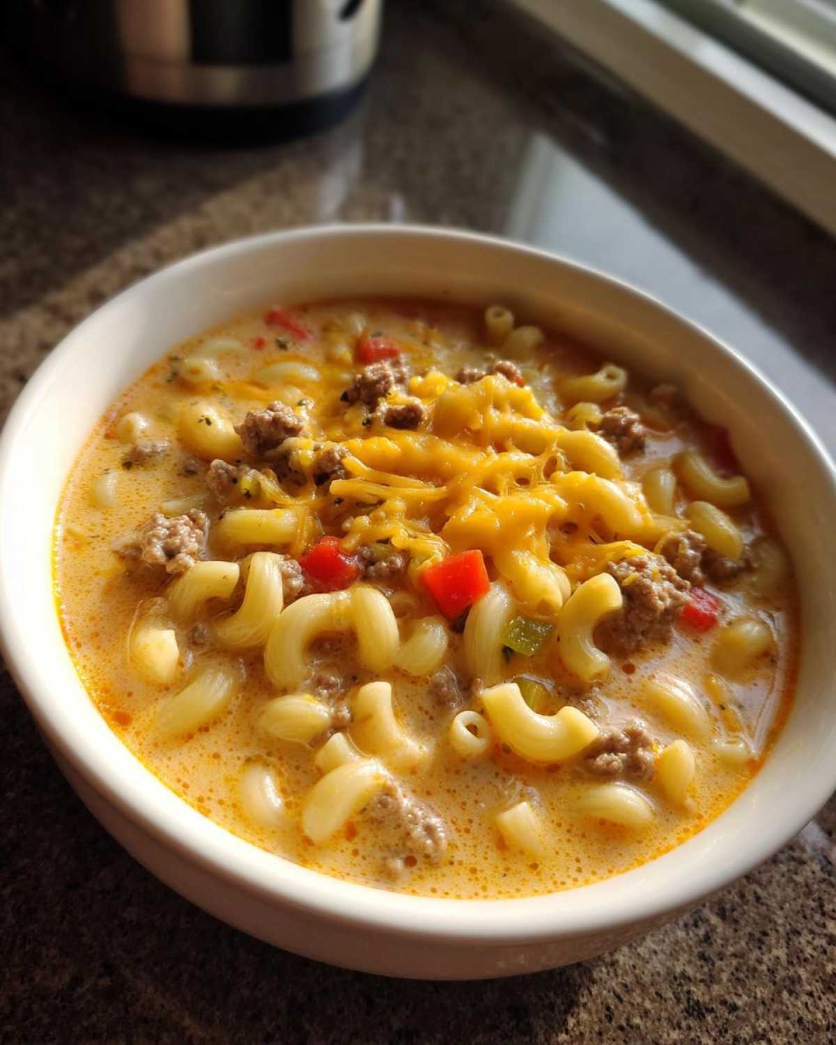 A close-up of a bowl of macaroni cheeseburger soup, featuring elbow macaroni, ground beef, and melted cheddar cheese.