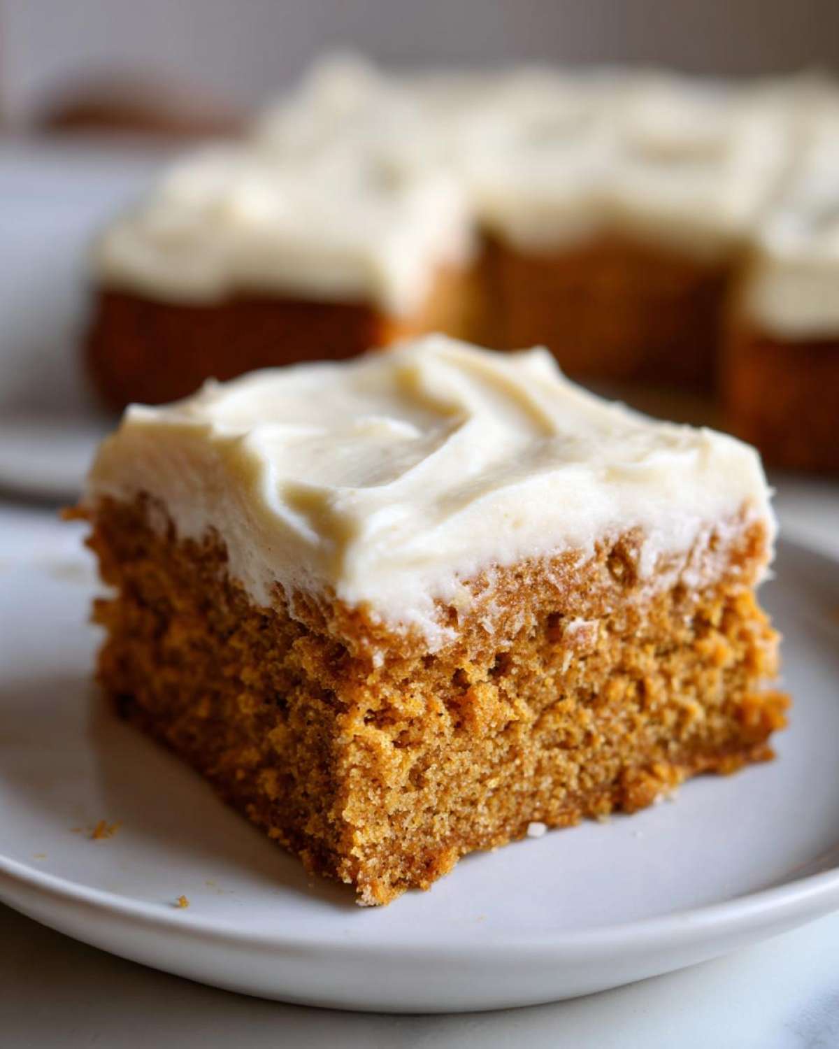 A slice of moist pumpkin bars topped with creamy frosting on a white plate.