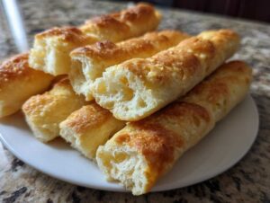A close-up of golden brown, soft Olive Garden breadsticks piled on a white plate, with some broken open to show the fluffy interior.