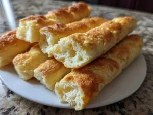 A close-up of golden brown, soft Olive Garden breadsticks piled on a white plate, with some broken open to show the fluffy interior.