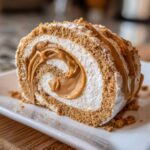 A slice of potato candy rolled cake, filled with whipped cream and caramel, topped with cookie crumbs and caramel drizzle.