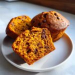 Three pumpkin banana muffins, one cut in half to show the texture and chocolate chips, on a white plate.