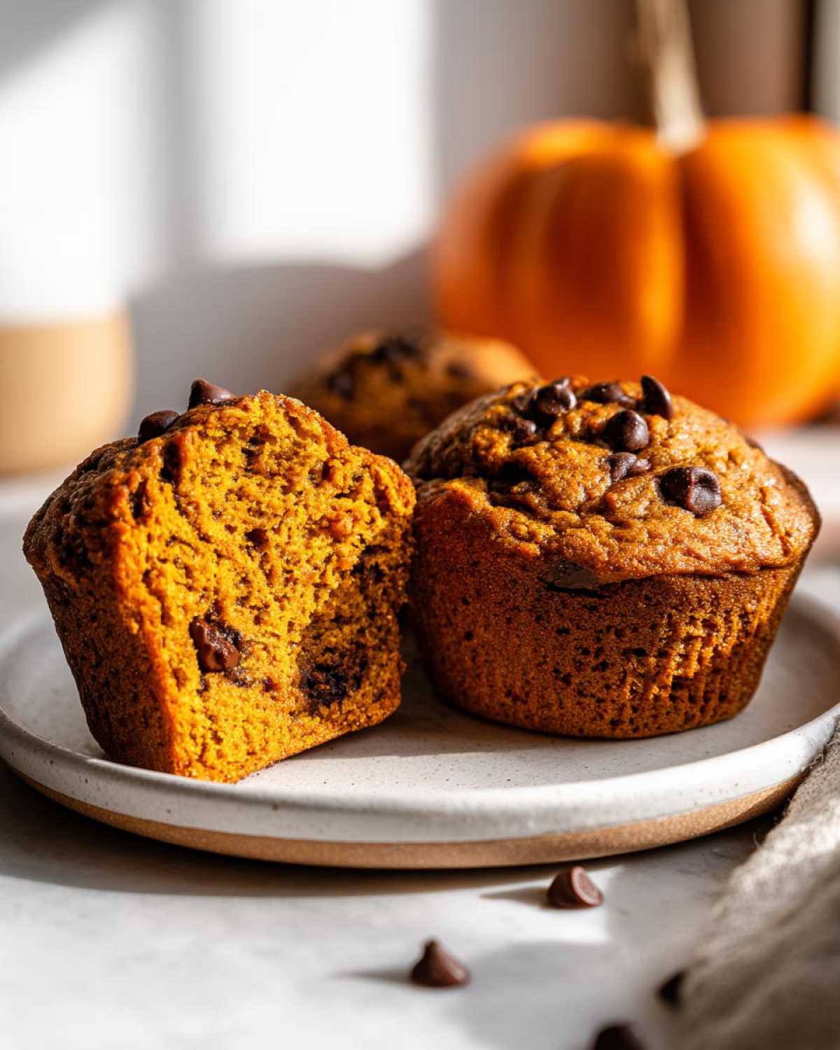 Two pumpkin banana muffins on a plate, one sliced to show the inside, topped with chocolate chips.