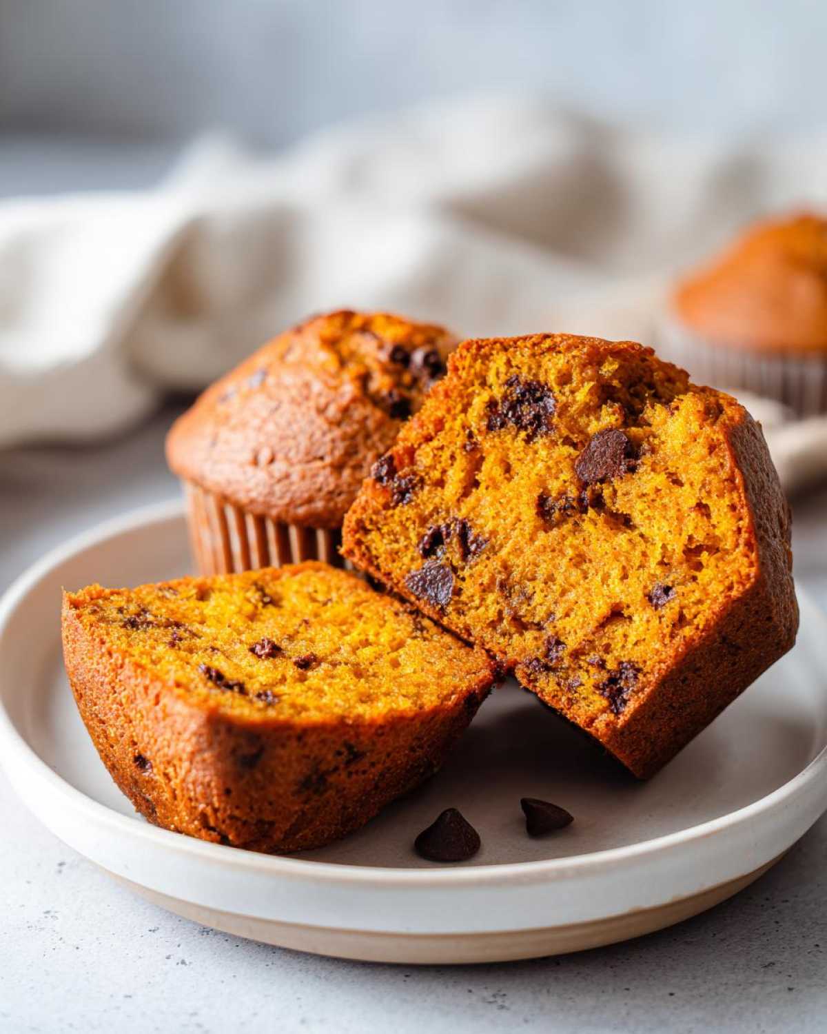 Close-up of pumpkin banana muffins on a plate, one muffin is cut in half to show the texture and chocolate chips.