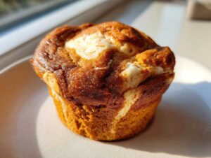 Close-up of a single pumpkin cheesecake muffin with a cream cheese swirl on a white plate.