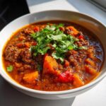 A close-up of a bowl of hearty pumpkin chili, featuring chunks of pumpkin, ground meat, and red peppers, topped with fresh cilantro.