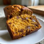 A slice of moist pumpkin chocolate chip bread on a white plate, with the loaf in the background.