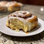 Close-up of a glazed pumpkin cinnamon roll on a white plate, with a bite taken out, revealing the soft interior.