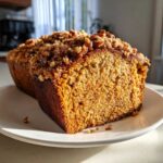 A slice of pumpkin loaf with a streusel topping on a white plate, showcasing the moist texture.