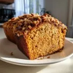 A slice of pumpkin loaf with a streusel topping on a white plate, showcasing the moist texture.
