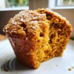 Close-up of a moist pumpkin muffin with a bite taken out, sprinkled with sugar crystals on a white plate.
