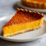 A close-up of a slice of creamy pumpkin pie recipe on a white plate, with a golden crust.