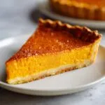 A close-up of a slice of creamy pumpkin pie recipe on a white plate, with a golden crust.