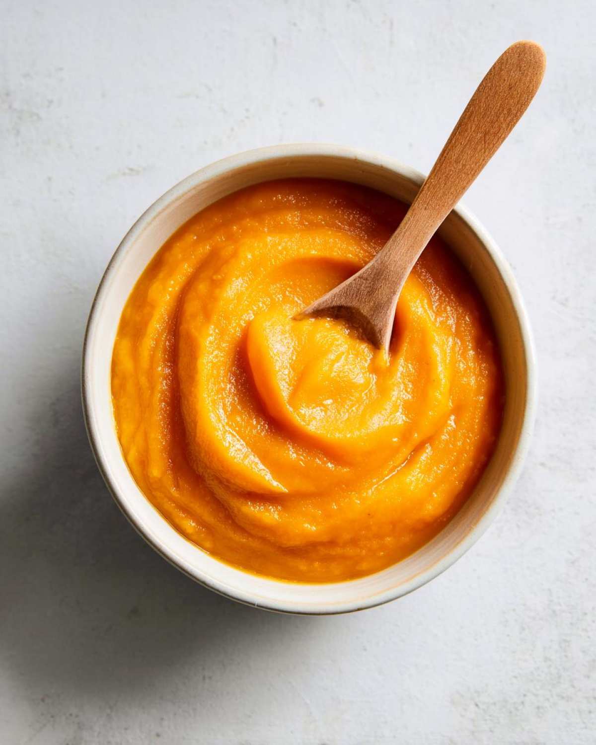 A close-up overhead shot of smooth, vibrant orange pumpkin puree in a bowl with a wooden spoon.