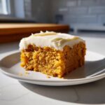 A close-up of a moist slice of pumpkin sheet cake topped with creamy white frosting on a white plate.