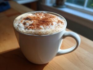 A white mug filled with a frothy pumpkin spice latte, topped with pumpkin spice, sits on a wooden surface.