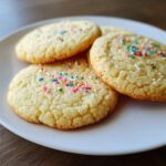 Close-up of three perfect sugar cookies topped with colorful sprinkles on a white plate.