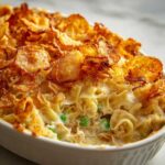 Close-up of creamy tuna casserole with potato chips topping, noodles, and peas in a white baking dish.