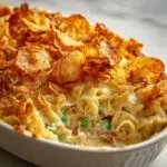Close-up of creamy tuna casserole with potato chips topping, noodles, and peas in a white baking dish.