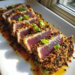 Slices of tuna tataki with a sesame seed crust, drizzled with sauce and garnished with green onions.