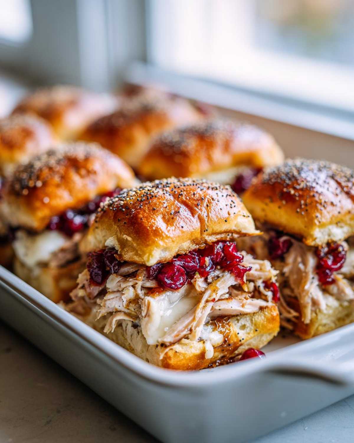 A close-up view of several turkey cranberry sliders in a white baking dish, topped with poppy seeds.