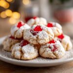 A close-up of a stack of whipped shortbread cookies, dusted with powdered sugar and topped with bright red cherries.