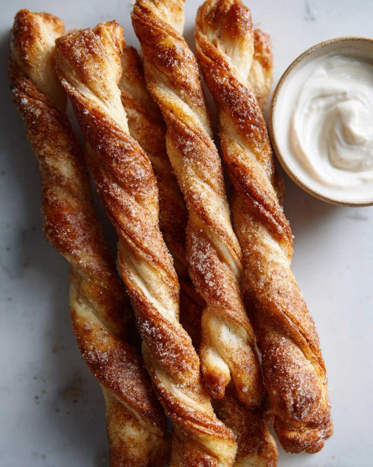 Close-up of golden brown air fryer cinnamon twists sprinkled with sugar and cinnamon, next to a small bowl of creamy dip.