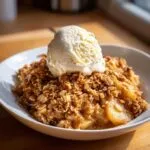A close-up of warm apple crisp with oat streusel topped with a scoop of vanilla ice cream in a white bowl.