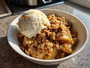 A close-up of a bowl of warm apple crisp with oat streusel, topped with a scoop of vanilla ice cream.