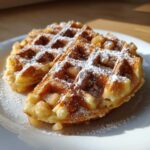 Close-up of two golden apple fritter waffle donuts dusted with powdered sugar and visible apple pieces.