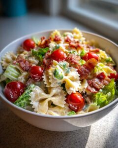 A close-up of a bowl of BLT pasta salad featuring farfalle pasta, cherry tomatoes, crisp lettuce, and crumbled bacon in a creamy dressing.