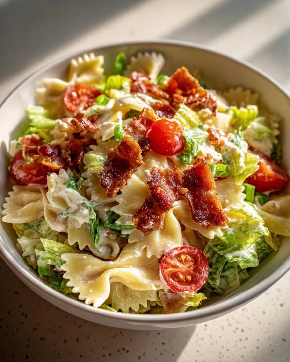 A close-up of a bowl of BLT pasta salad featuring farfalle pasta, crispy bacon, cherry tomatoes, and lettuce.