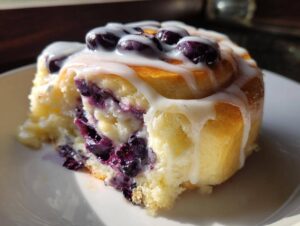 A close-up of a delicious blueberry cheesecake roll, drizzled with white icing and topped with fresh blueberries.