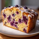 A close-up of a slice of blueberry loaf cake, drizzled with white glaze and topped with fresh blueberries.