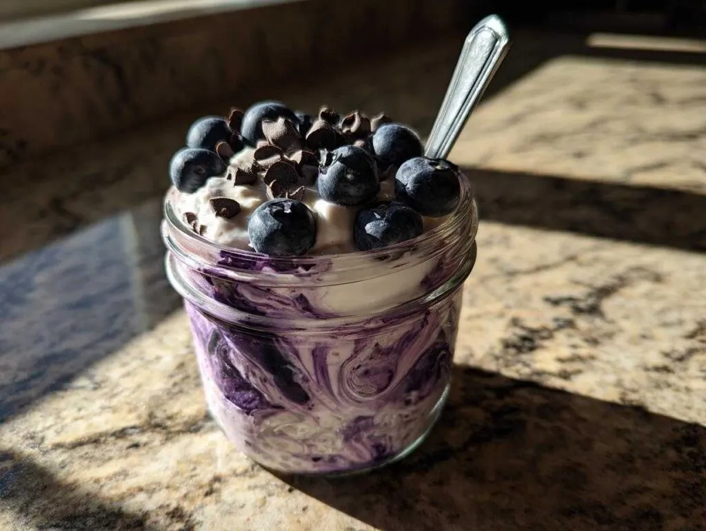 A close-up of creamy blueberry overnight oats in a jar, topped with fresh blueberries and chocolate chips, with a spoon.