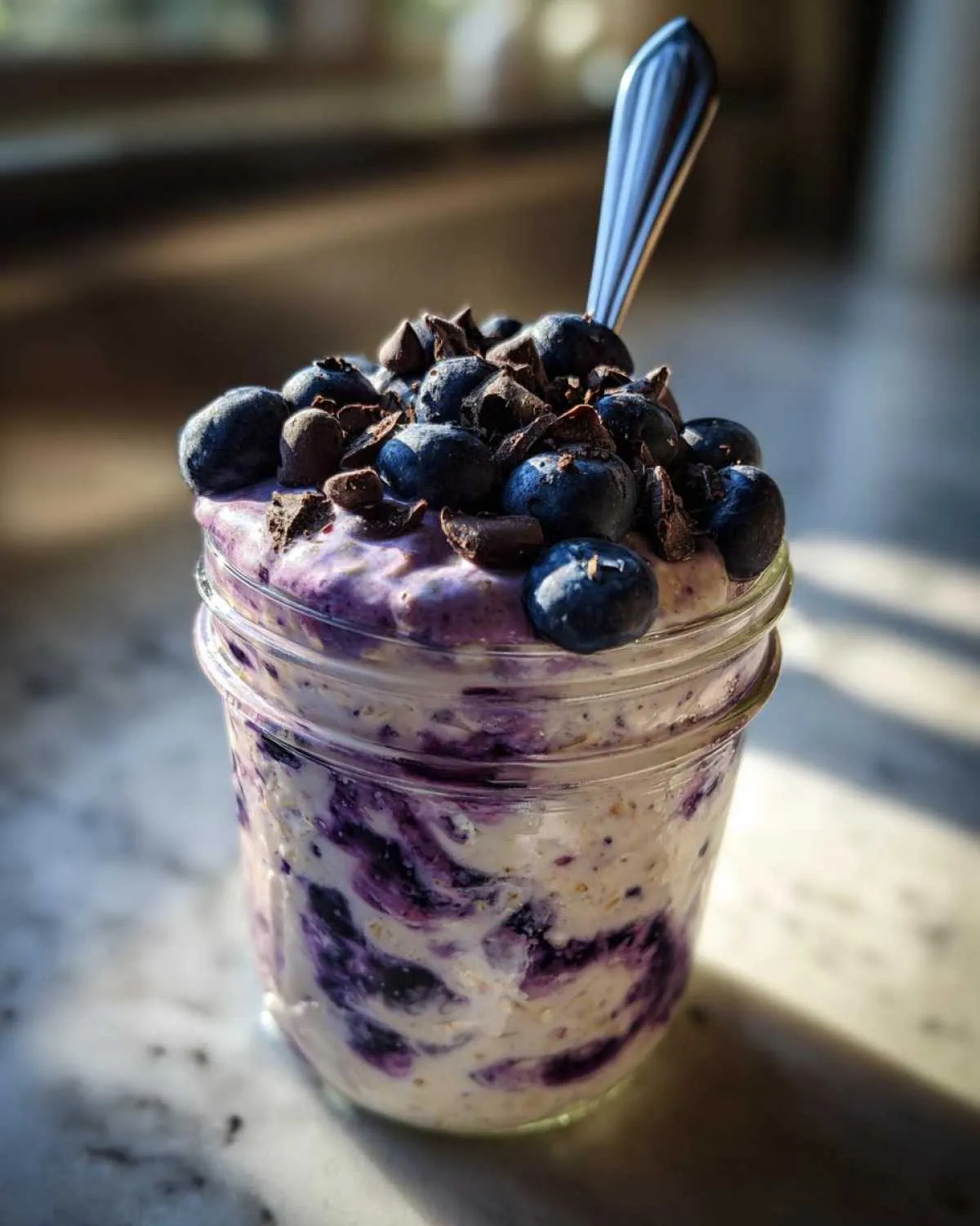 A close-up of creamy blueberry overnight oats in a jar, topped with fresh blueberries and chocolate chips, with a spoon.