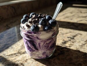 A close-up of creamy blueberry overnight oats in a jar, topped with fresh blueberries and chocolate chips, with a spoon.