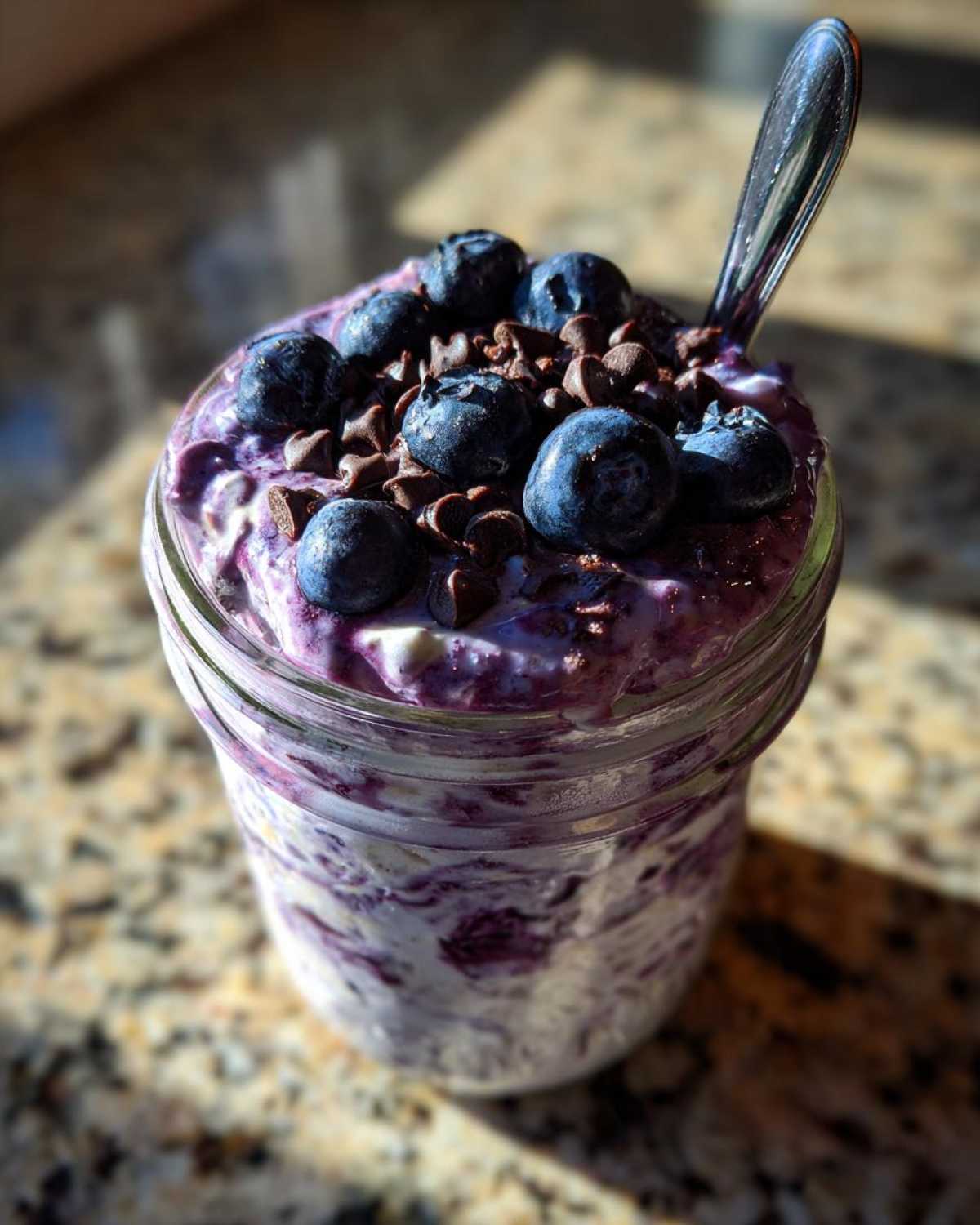 Close-up of blueberry overnight oats in a jar, topped with fresh blueberries and chocolate chips, with a spoon.