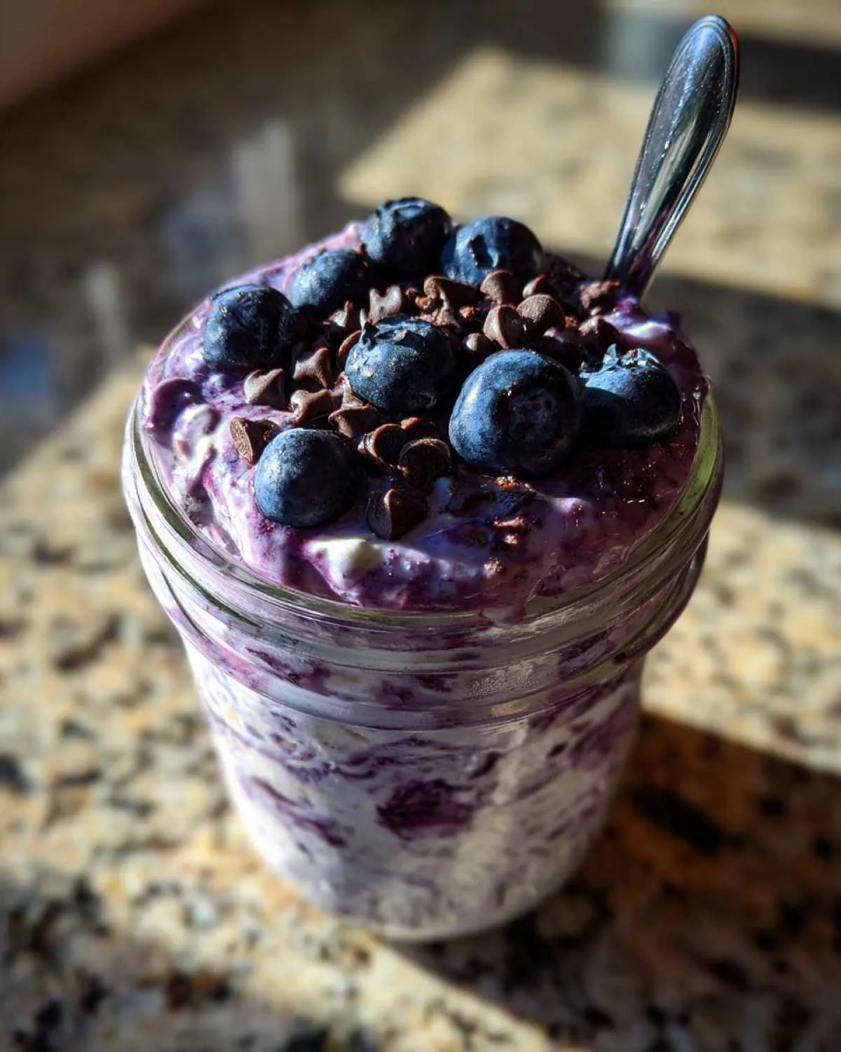 Close-up of blueberry overnight oats in a jar, topped with fresh blueberries and chocolate chips, with a spoon.